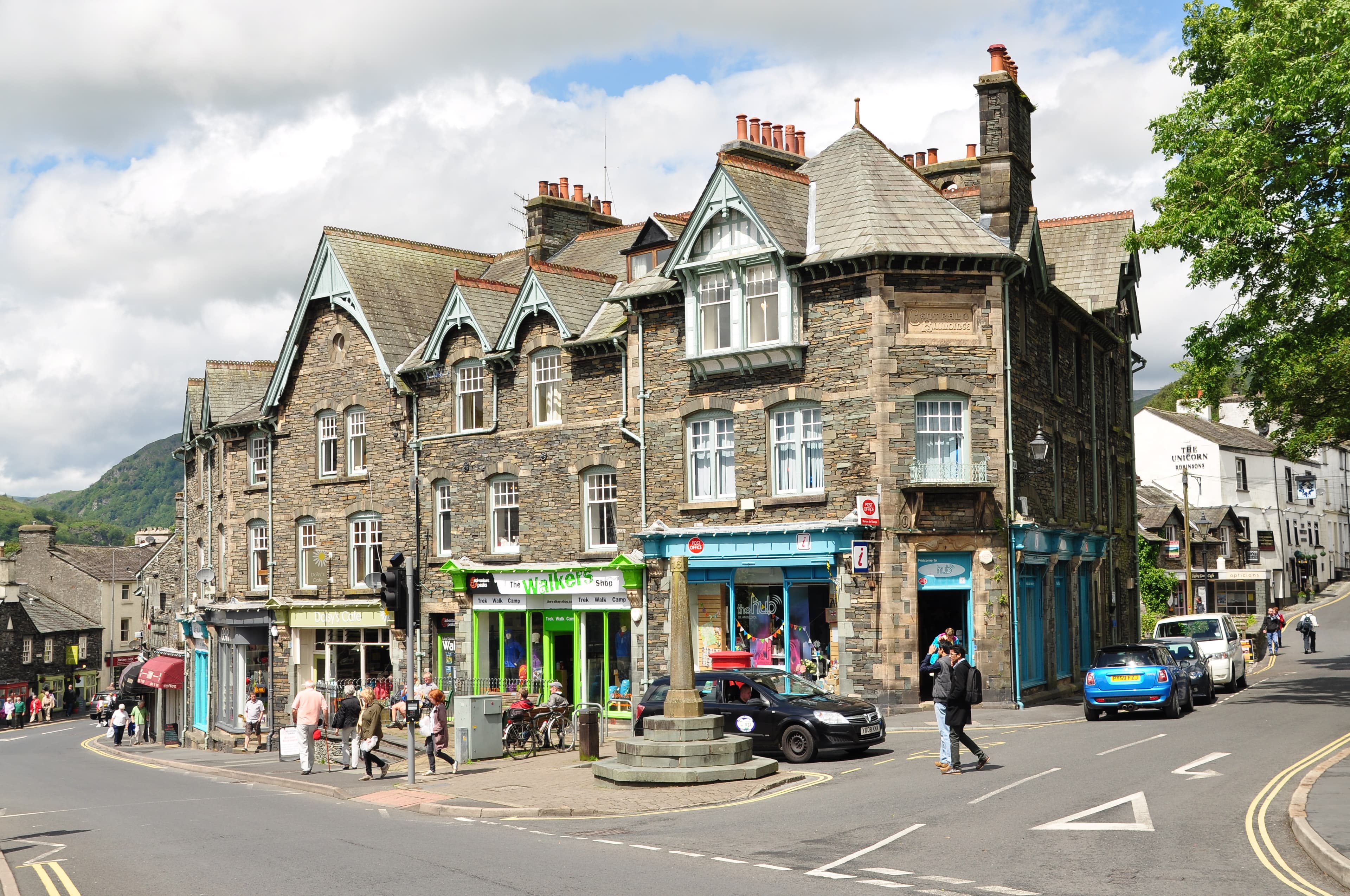 Slate and stone buildings on the corner of the main street in Ambleside, with shops, a market cross and Lakeland fells rising behind.