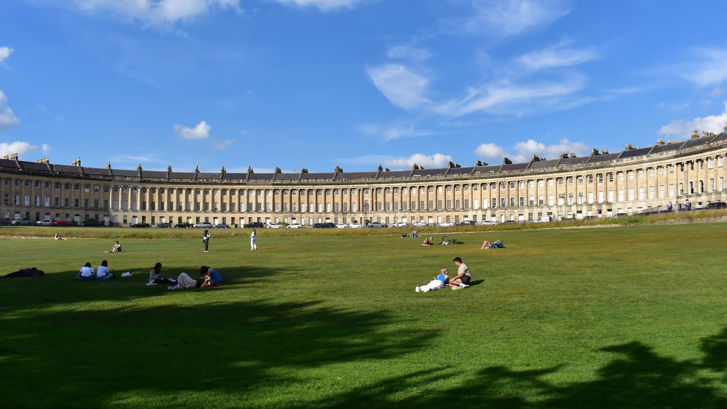 The curved Georgian terrace of the Royal Crescent in Bath, seen across the open lawn where people sit in the sun.