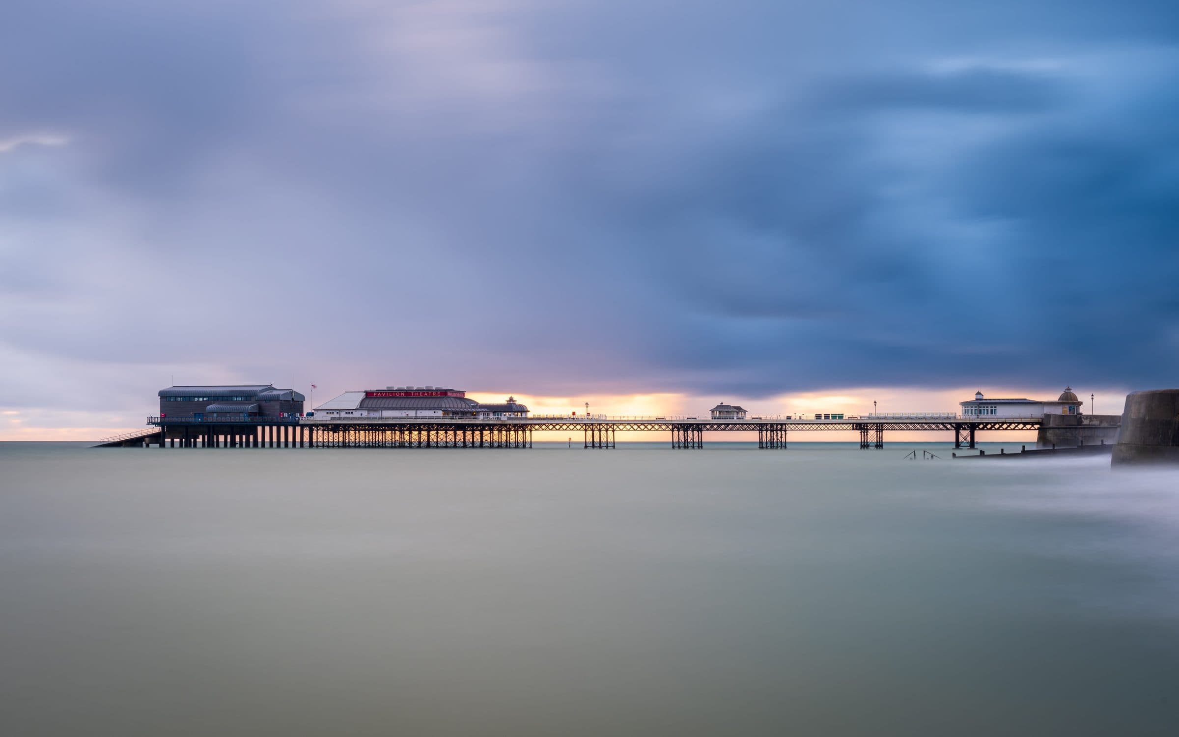 Cromer Pier reaching out into a calm sea at dusk, its lifeboat station and pavilion theatre silhouetted against the sky.